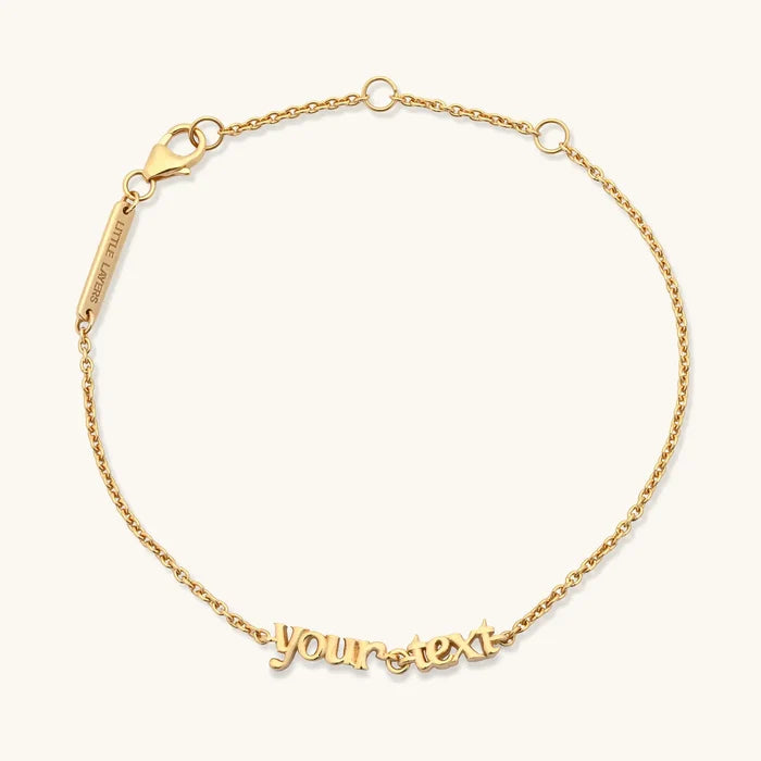 A delicate gold chain bracelet with a personalized script nameplate reading 'your text' and a lobster clasp, shown on a light cream background.