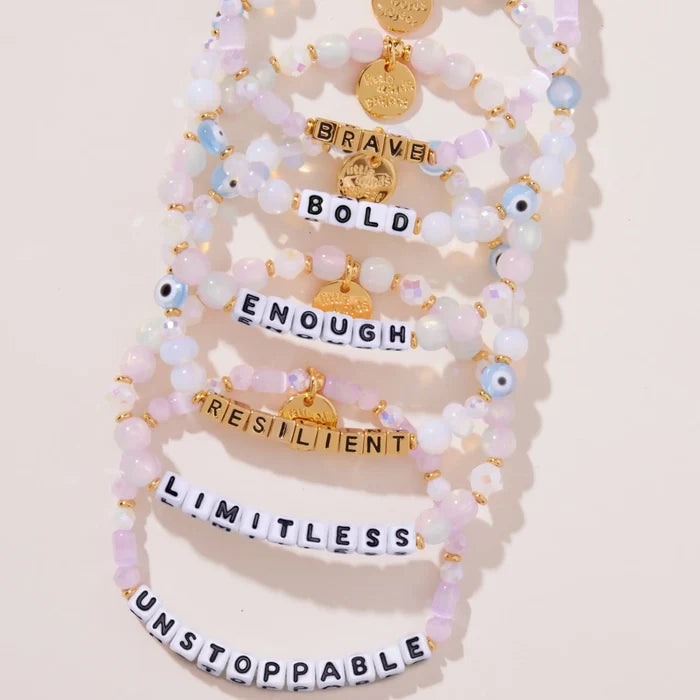 Six Mystic Blessings beaded bracelets laid flat in a stack reading BRAVE, BOLD, ENOUGH, RESILIENT, LIMITLESS, and UNSTOPPABLE, pastel pink and iridescent beads with gold hardware