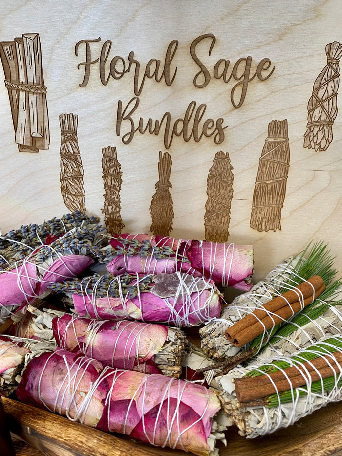 Hand-wrapped floral sage bundles with rose petals and cinnamon in front of an engraved Floral Sage Bundles sign