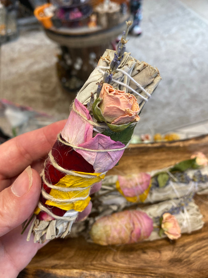 White sage smudge stick wrapped with dried roses and petals held in hand; each stick's florals vary