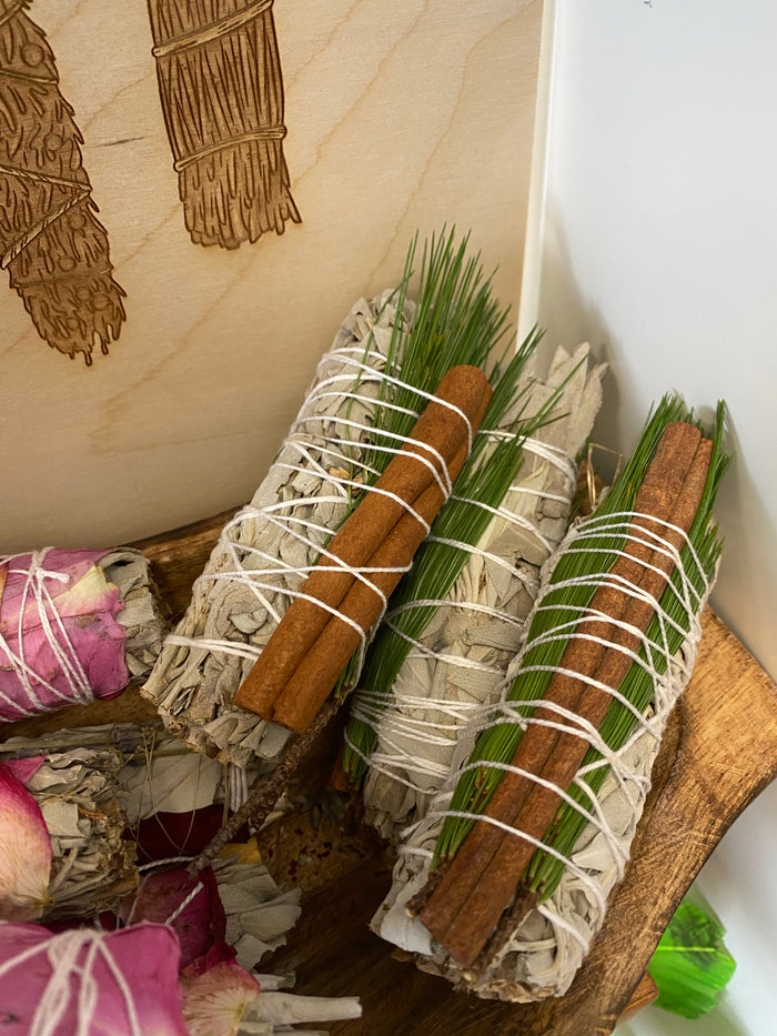 White sage smudge sticks wrapped with cinnamon sticks and green sprigs in a wooden bowl