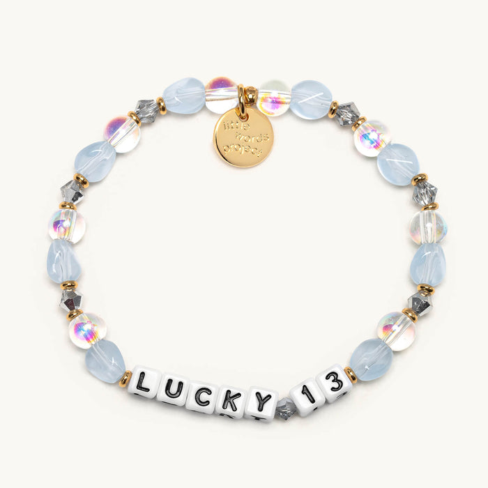 Lucky 13 bracelet flat-lay with frosted light-blue and iridescent clear beads, silver crystal accents, gold spacers, and a gold Little Words Project charm