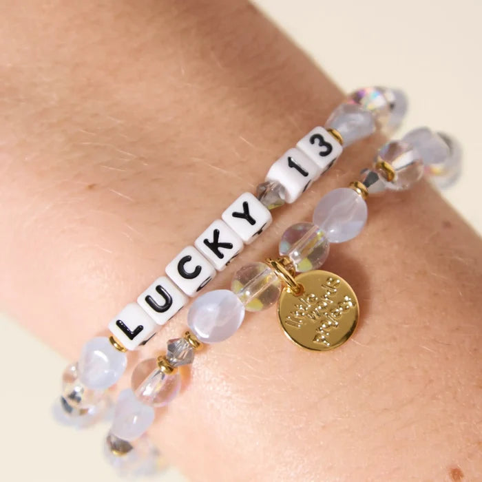 Two Lucky 13 bracelets stacked on a wrist, with frosted blue and iridescent beads, a gold Little Words Project charm, and white LUCKY 13 letter beads