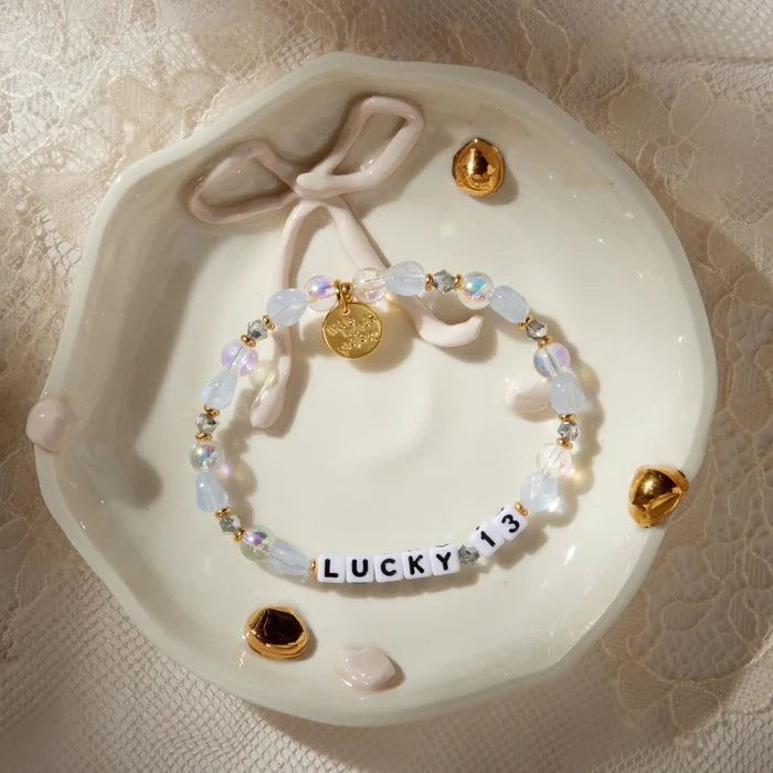 Lucky 13 bracelet styled in a white ceramic trinket dish with a bow detail and gold accents on lace, frosted blue and iridescent beads
