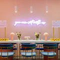 Modern interior with a long table, blue chairs, and decorative elements against a pink wall with a neon sign.