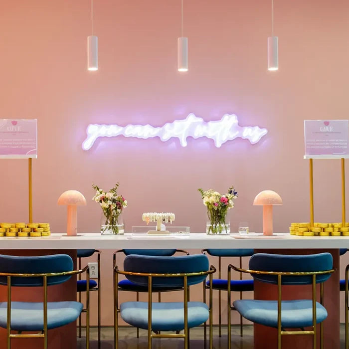 Modern interior with a long table, blue chairs, and decorative elements against a pink wall with a neon sign.